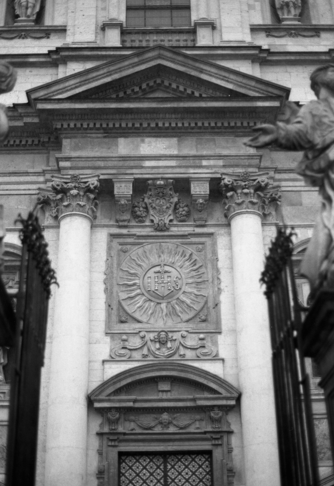 Barokowa fasada kościoła z płaskorzeźbą słońca z monogramem, między kolumnami i kutą bramą