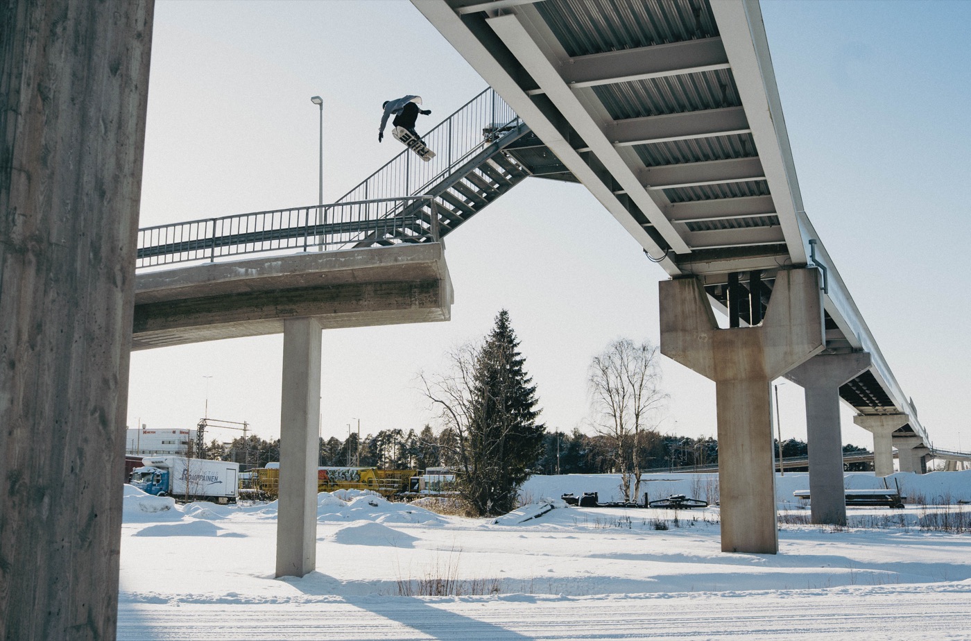 Snowboardzista w powietrzu nad schodami kładki, w tle zaśnieżone miasto