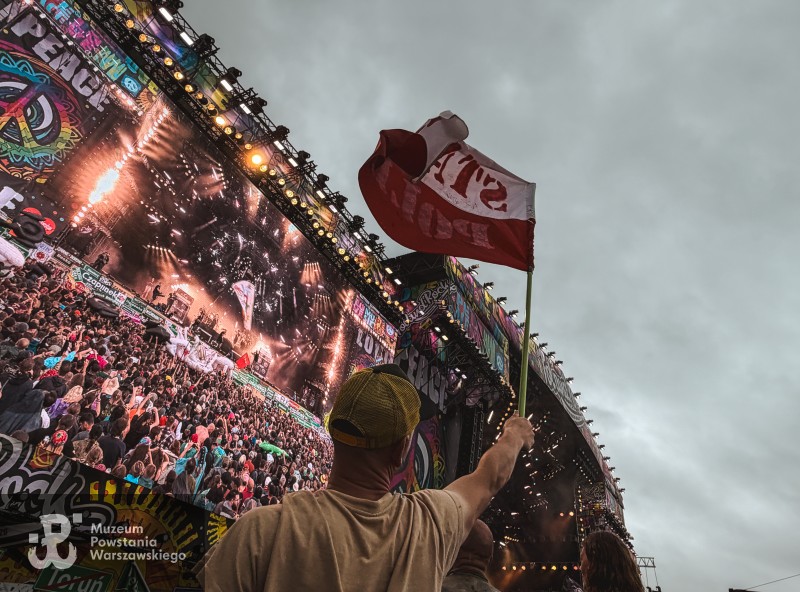 Mężczyzna unosi małą flagę przed oświetloną sceną i tłumem