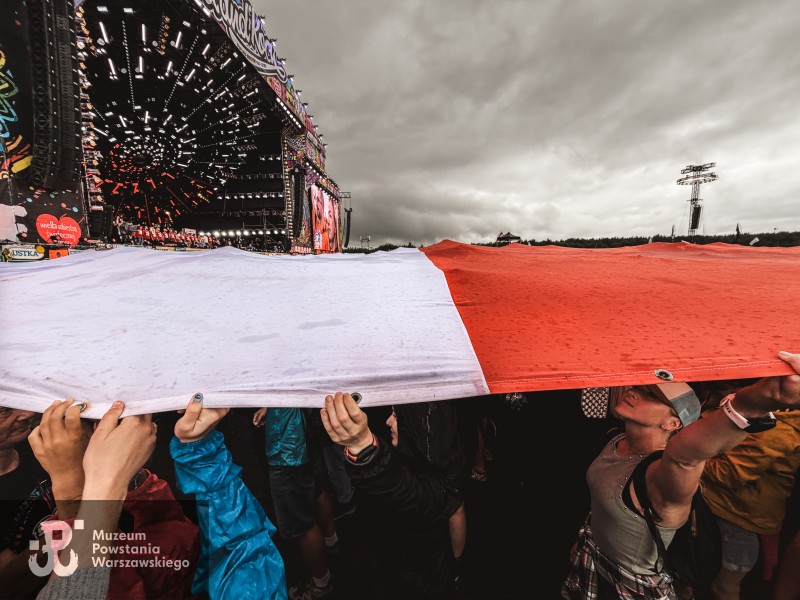 Tłum trzyma nad głowami ogromną biało-czerwoną flagę przed sceną
