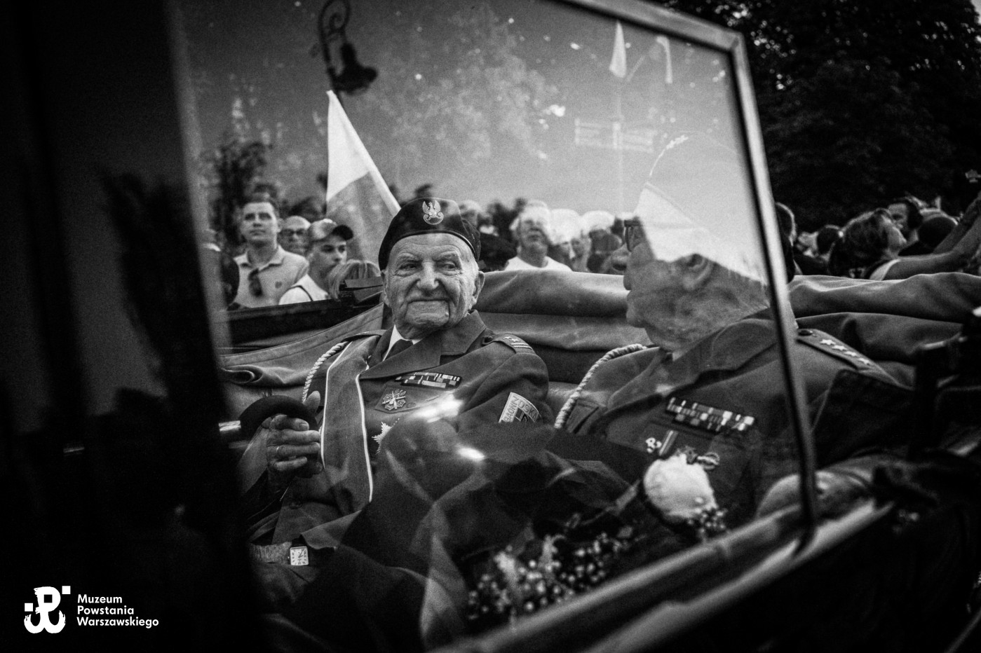Czarno-białe zdjęcie starszego mężczyzny w mundurze i berecie w samochodzie, w szybie odbija się tłum z flagą