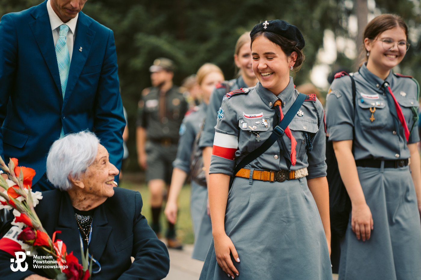 Starsza kobieta z kwiatami uśmiecha się do przechodzących harcerek