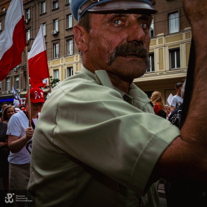 Mężczyzna z wąsem w czapce z daszkiem i koszuli w kolorze khaki, w tle flagi i kamienice