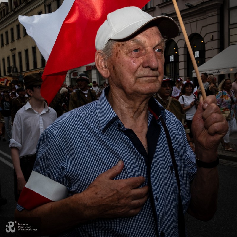 Starszy mężczyzna z biało-czerwoną opaską trzyma flagę i kładzie dłoń na piersi